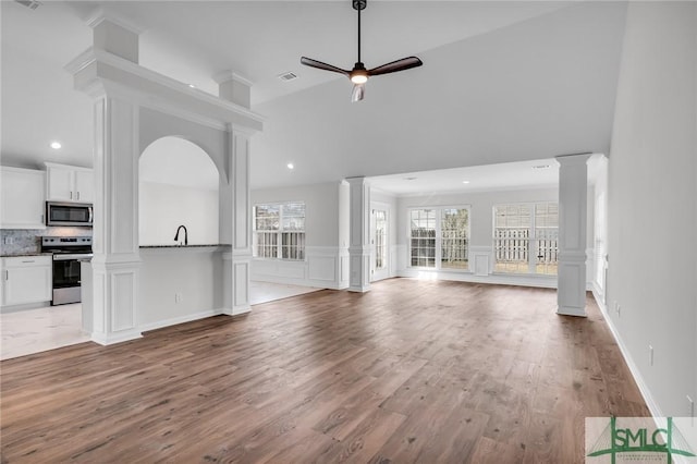 unfurnished living room featuring ceiling fan, a towering ceiling, light wood-type flooring, and ornate columns