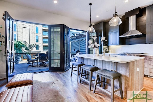 kitchen featuring sink, light hardwood / wood-style flooring, pendant lighting, a kitchen island with sink, and wall chimney range hood