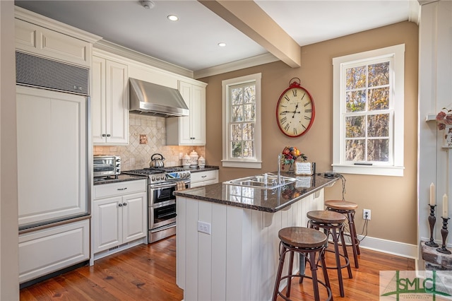 kitchen featuring wall chimney range hood, a breakfast bar area, dark stone countertops, double oven range, and tasteful backsplash