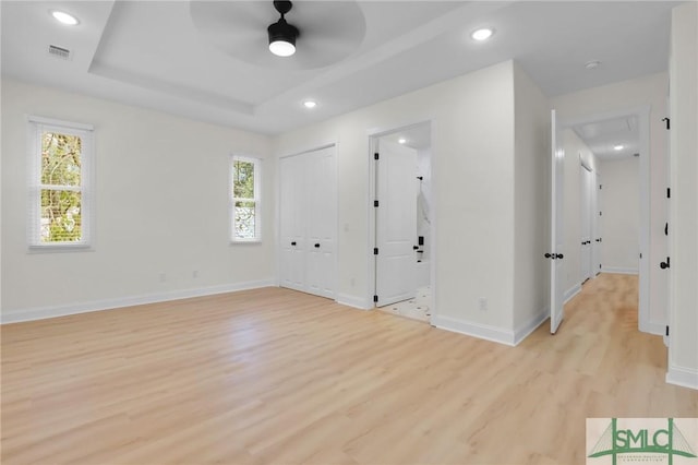 empty room with ceiling fan, a tray ceiling, light wood-type flooring, and a wealth of natural light