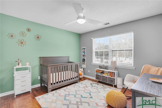 bedroom with a crib, ceiling fan, a textured ceiling, and dark hardwood / wood-style flooring