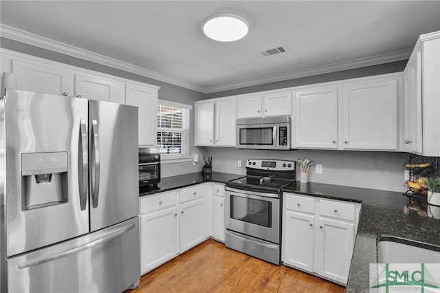 kitchen with appliances with stainless steel finishes, white cabinets, dark stone counters, crown molding, and light wood-type flooring