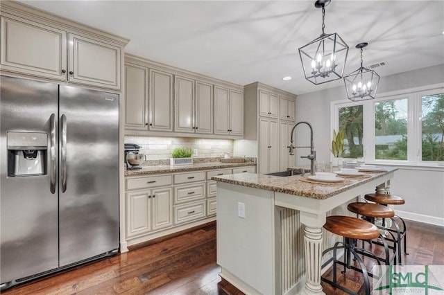 kitchen with sink, stainless steel refrigerator with ice dispenser, light stone countertops, an island with sink, and cream cabinetry