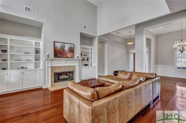 living room featuring a towering ceiling, built in shelves, a tile fireplace, an inviting chandelier, and dark hardwood / wood-style floors