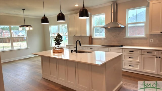 kitchen with a sink, hanging light fixtures, wall chimney range hood, light countertops, and a center island with sink