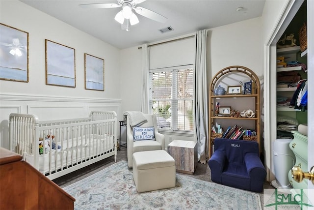 bedroom featuring a crib, visible vents, wainscoting, ceiling fan, and a decorative wall