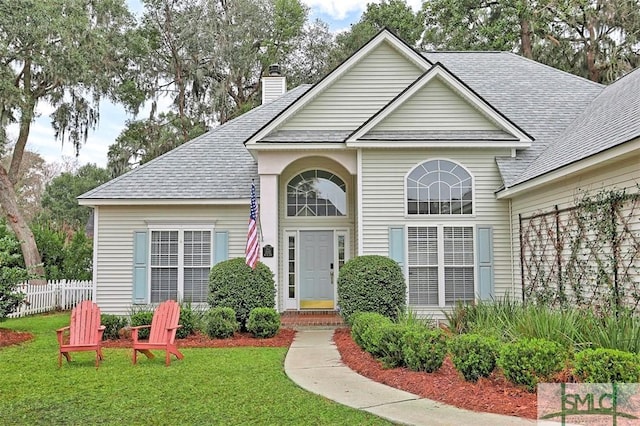 traditional-style house featuring roof with shingles, fence, a chimney, and a front lawn