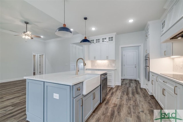 kitchen with glass insert cabinets, appliances with stainless steel finishes, a kitchen island with sink, light countertops, and white cabinetry