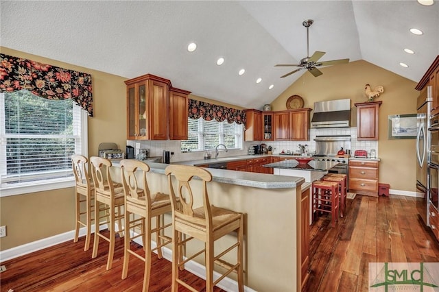 kitchen featuring a peninsula, a kitchen breakfast bar, wall chimney exhaust hood, brown cabinetry, and glass insert cabinets