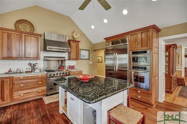 kitchen with a kitchen island, high quality appliances, wall chimney range hood, brown cabinetry, and dark stone countertops