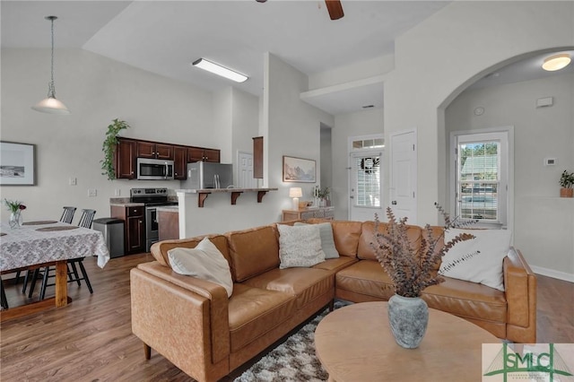 living room featuring arched walkways, high vaulted ceiling, light wood-style flooring, a ceiling fan, and baseboards