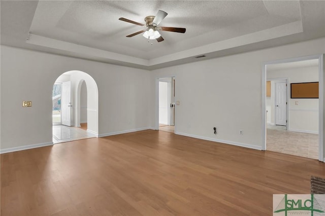 empty room with arched walkways, a raised ceiling, ceiling fan, a textured ceiling, and wood finished floors