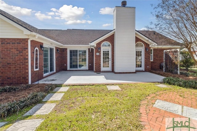 back of house with brick siding, roof with shingles, a lawn, a chimney, and a patio area