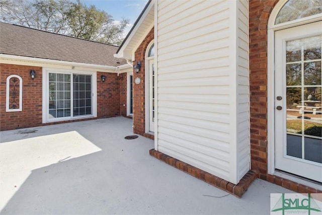 view of exterior entry featuring a patio area, a shingled roof, and brick siding