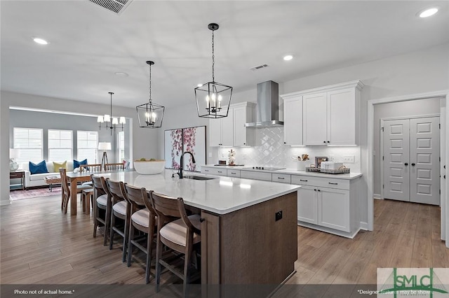 kitchen featuring light wood finished floors, wall chimney exhaust hood, white cabinets, a sink, and a kitchen bar