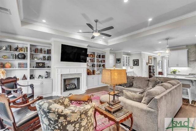 living room with a ceiling fan, a fireplace with flush hearth, wood finished floors, a tray ceiling, and crown molding