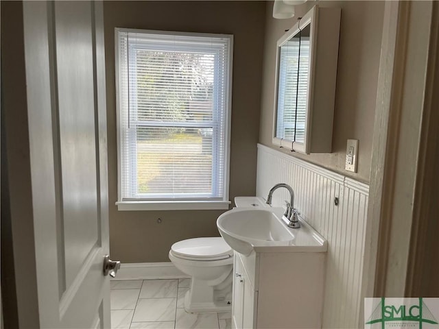 half bathroom with marble finish floor, toilet, wainscoting, vanity, and baseboards