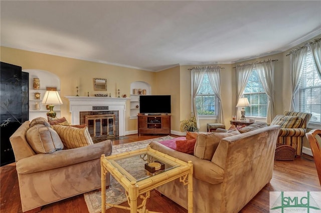 living area with built in shelves, wood finished floors, a fireplace with flush hearth, baseboards, and crown molding