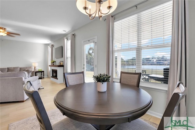 dining space with light wood-style floors, a glass covered fireplace, baseboards, and ceiling fan with notable chandelier