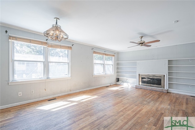 unfurnished living room with ceiling fan with notable chandelier, baseboards, ornamental molding, hardwood / wood-style floors, and a glass covered fireplace