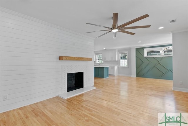 unfurnished living room with visible vents, a fireplace with raised hearth, ceiling fan, crown molding, and light wood-style floors