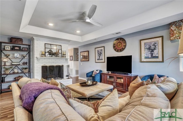 living area featuring wood finished floors, visible vents, a ceiling fan, a brick fireplace, and a raised ceiling