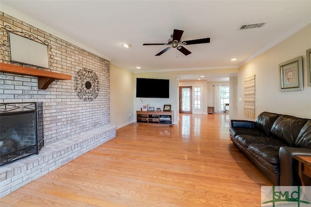 living room with crown molding, visible vents, a ceiling fan, a brick fireplace, and light wood-type flooring