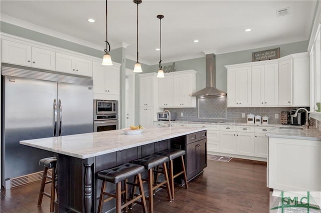 kitchen featuring visible vents, built in appliances, wall chimney range hood, white cabinetry, and a sink