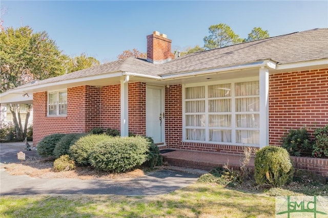 property entrance with an attached carport, brick siding, a chimney, and roof with shingles
