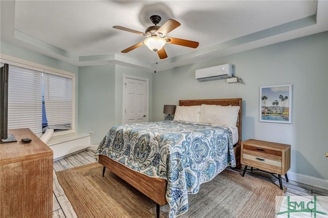 bedroom featuring a wall mounted air conditioner, a raised ceiling, wood finished floors, baseboards, and ceiling fan