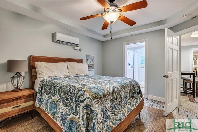 bedroom featuring wood finished floors, a wall unit AC, connected bathroom, baseboards, and ceiling fan