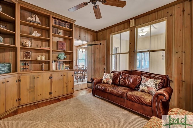 living area featuring light wood-style flooring, crown molding, wooden walls, and ceiling fan with notable chandelier