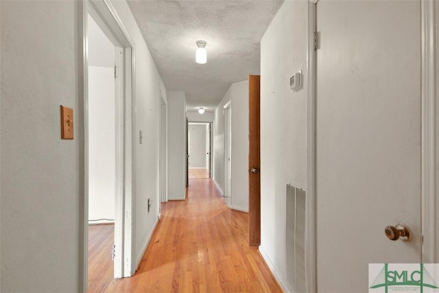 hall featuring light wood-type flooring, visible vents, a textured ceiling, and baseboards