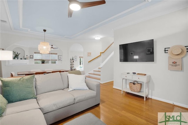 living area featuring arched walkways, ceiling fan, stairway, light wood-type flooring, and crown molding