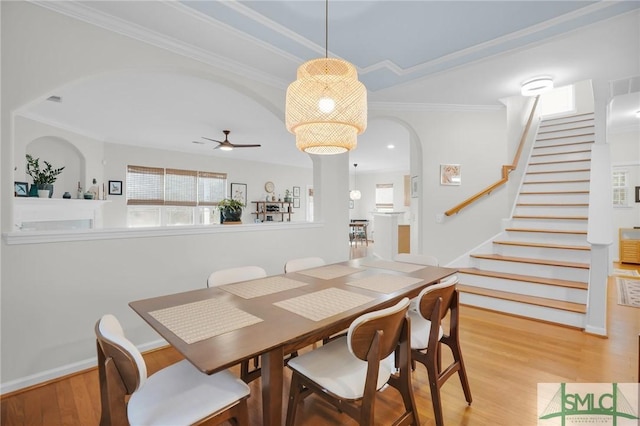 dining area featuring arched walkways, light wood-style flooring, ornamental molding, ceiling fan, and stairs