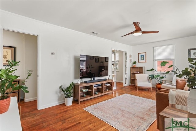 living area featuring wood finished floors, visible vents, baseboards, a ceiling fan, and crown molding