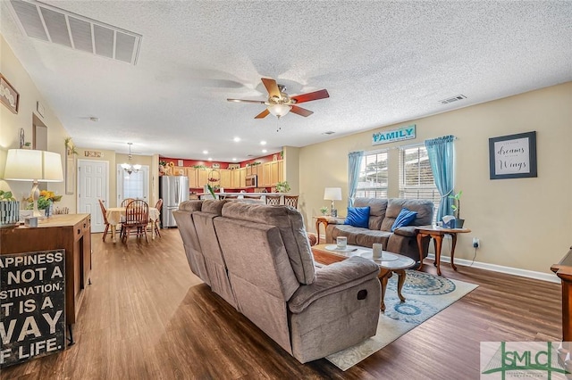 living area featuring a textured ceiling, ceiling fan with notable chandelier, wood finished floors, visible vents, and baseboards