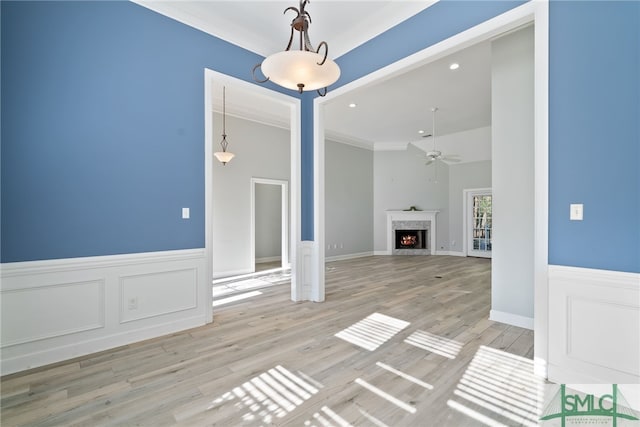 unfurnished living room with ceiling fan, a wainscoted wall, a lit fireplace, ornamental molding, and light wood finished floors