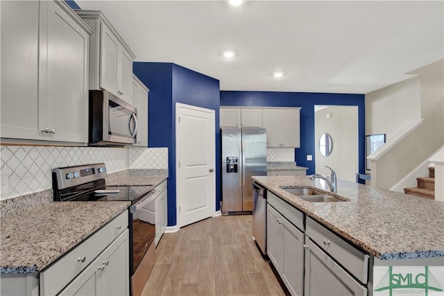 kitchen with stainless steel appliances, light wood-style flooring, a kitchen island with sink, a sink, and light stone countertops