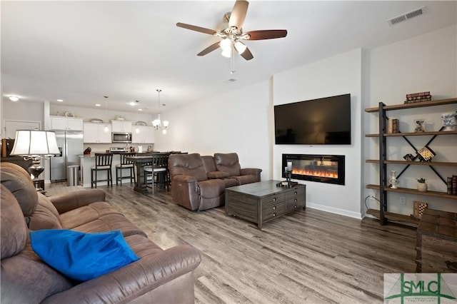 living room with visible vents, a glass covered fireplace, wood finished floors, baseboards, and ceiling fan with notable chandelier