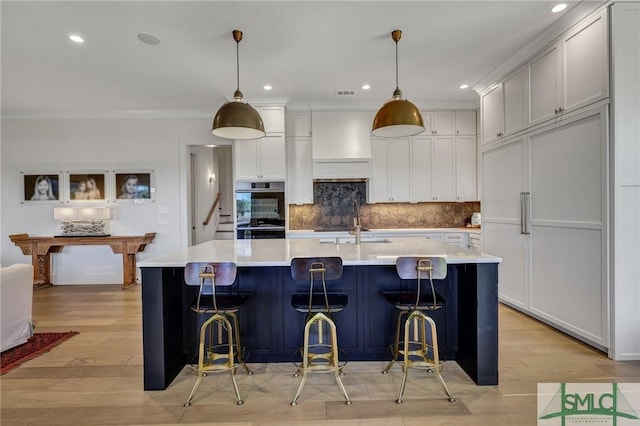 kitchen featuring light wood-style flooring, light countertops, a large island with sink, double oven, and backsplash