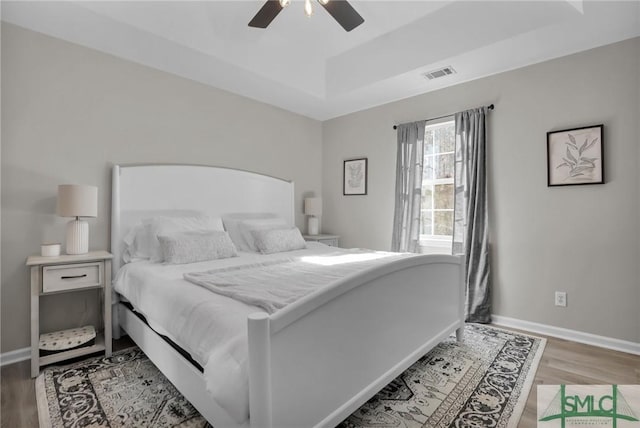 bedroom with a raised ceiling, visible vents, baseboards, and wood finished floors