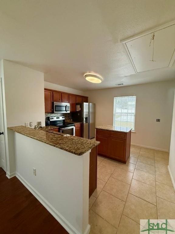 kitchen with light tile patterned floors, appliances with stainless steel finishes, brown cabinetry, a peninsula, and baseboards