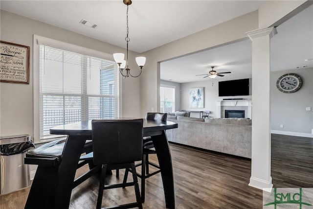 dining area featuring ceiling fan with notable chandelier, dark wood finished floors, visible vents, a glass covered fireplace, and ornate columns