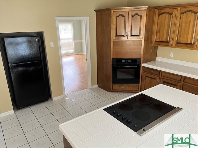 kitchen featuring black appliances, baseboards, tile counters, and brown cabinetry