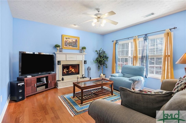 living room with a textured ceiling, ceiling fan, wood finished floors, visible vents, and a lit fireplace