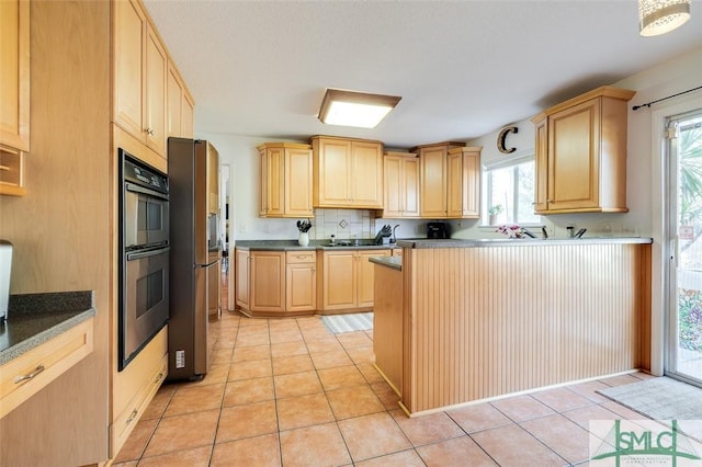 kitchen with light tile patterned floors, dark countertops, stainless steel refrigerator, double wall oven, and light brown cabinetry