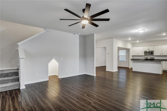 unfurnished living room with stairs, ceiling fan, dark wood-type flooring, and baseboards