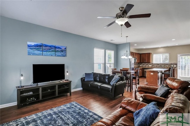 living area featuring dark wood finished floors, plenty of natural light, ceiling fan with notable chandelier, and baseboards