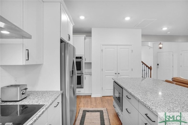 kitchen featuring light stone countertops, light wood-type flooring, appliances with stainless steel finishes, white cabinets, and wall chimney exhaust hood